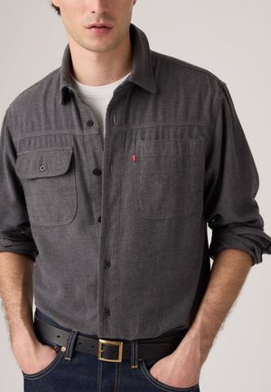 Gray button-up shirt made of textured fabric, featuring two chest pockets, and a small red logo, worn with a black belt and dark jeans.
