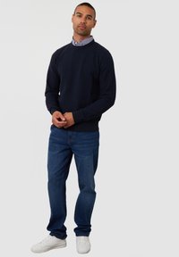 Marineblauwe gebreide trui met geribde manchetten en zoom, gecombineerd met blauwe jeans en witte sneakers. Model staat tegen een effen achtergrond.