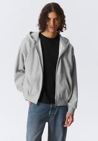 Jeune personne aux cheveux bouclés portant un sweat à capuche gris zippé sur un t-shirt noir et un jean bleu, debout devant un fond blanc.