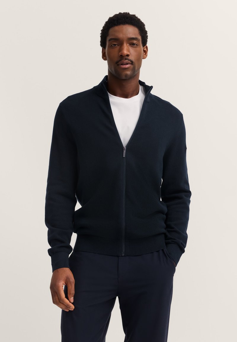 Uomo che indossa un maglione blu navy con zip sopra una camicia bianca e pantaloni scuri, in piedi con una mano in tasca su uno sfondo neutro.