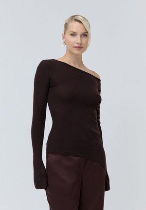 Femme aux cheveux blonds portant un pull côtelé sombre à une épaule et un pantalon sombre assorti, debout devant un fond clair uni.