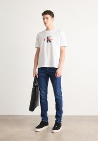 Hvit bomulls T-skjorte med svart og rød logo, kombinert med blå denimjeans. Svart veske holdt i hånden, avsluttet med svart og hvite joggesko.