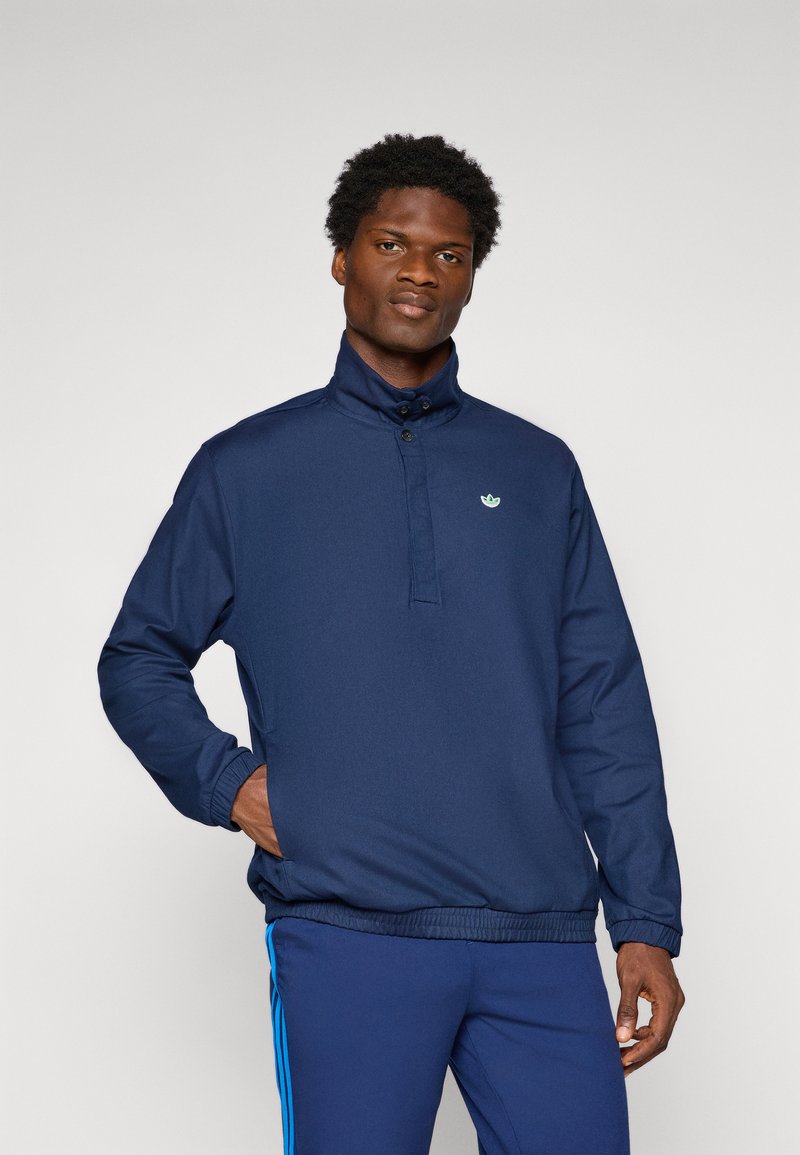 Jovem vestido com um pulôver azul-marinho com botões no colarinho alto, mãos nos bolsos, usando calças azuis-marinho combinando com riscas azul-claro.