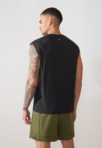 Juodas be rankovių sportinis marškinėlis su apvaliu iškirpimu, lygia tekstūra ir minimalizuotu dizainu. Dėvimi žali šortai. Matomi tatuiruotės detalės ant rankos.