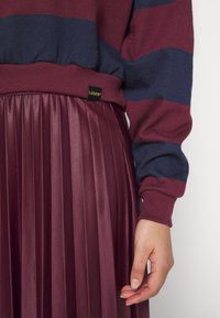 Personne portant un pull Levi’s rayé bordeaux et bleu marine avec une jupe plissée bordeaux, main droite détendue sur le côté sur un fond uni.