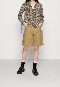 Blomstret bluse med V-hals, parret med khaki cargo shorts med sidelommer. Modellen har sorte snørebodder med kraftige såler på.
