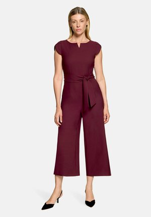 Kvinde iført en ærmeløs burgundy jumpsuit med bælte i taljen og brede cropped ben, parret med sorte spidse hæle mod en hvid baggrund.