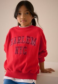 Rød sweatshirt med "HARLEM NYC" i fed skrift. Materiale i bomuld, rund hals, ribbede manchetter og kant. Laget over en hvid skjorte og blå bukser.