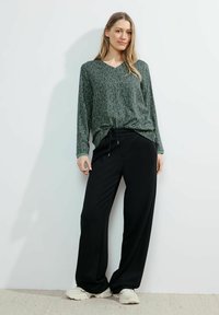 Top lungo a maniche lunghe a fantasia verde con scollatura a V, abbinato a pantaloni neri a gamba larga. Le scarpe sono di stile atletico di colore chiaro. Sfondo semplice.