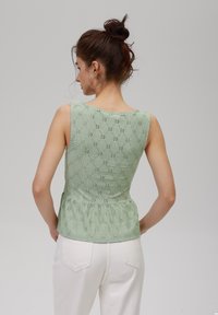 Gaiši zaļš peplum tops, izgatavots no teksturētas auduma, ar ziedu rakstu un bez piedurknēm, harmoniski apvienots ar baltām biksēm.