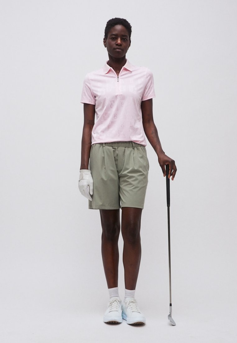 Femme portant un polo rose, un short vert, un gant de golf blanc et des baskets, debout avec un club de golf sur fond blanc.