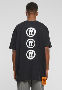 Melns kokvilnas T-krekls ar trim lielām baltām apļveida logo uz aizmugurējās daļas. Tai ir oranžs birka ar zīmola informāciju apakšējā daļā.