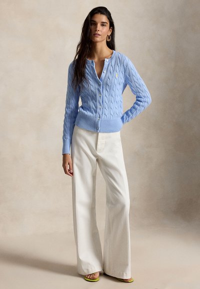 Nő világoskék kötött, gombos kardigánban, fehér, bő szárú nadrágban és zöld, nyitott orrú szandálban, egyszínű háttér előtt.