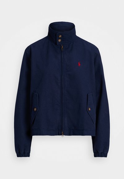 Casaco azul marinho com colarinho levantado, fecho de correr, dois bolsos laterais e detalhes em botões castanhos. Apresenta um pequeno logótipo vermelho no peito.