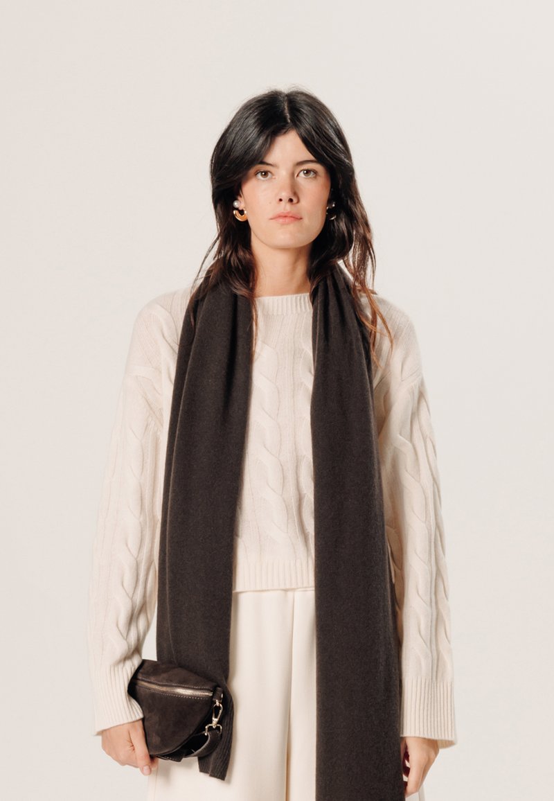 Kabelgebreide crèmekleurige trui gecombineerd met een lange, donkerbruine sjaal en een kleine, donkerleren pouch tas met rits. Eenvoudige, gelaagde look.