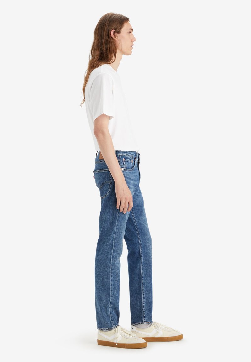 Weißes T-Shirt mit kurzen Ärmeln, blaue Denim-Jeans im geraden Schnitt und cremefarbene Sneakers mit strukturiertem Sohlen. Seitenansicht des Outfits.