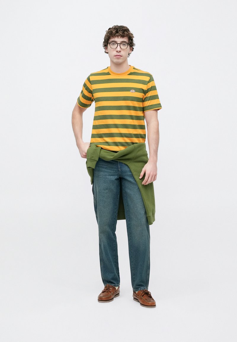 Maglietta a righe verdi e gialle con scollo rotondo, abbinata a jeans blu scuro e un maglione verde annodato in vita. Scarpe marroni.