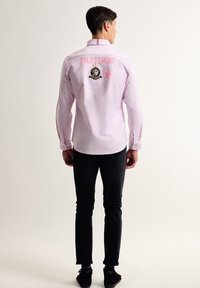 Camisa de manga larga rosa con el bordado "POLO LEGEND" y el logo en la parte trasera, combinada con pantalones negros y zapatos oscuros. Textura de tela suave.