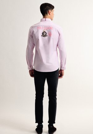 Camisa de manga larga rosa con el bordado "POLO LEGEND" y el logo en la parte trasera, combinada con pantalones negros y zapatos oscuros. Textura de tela suave.