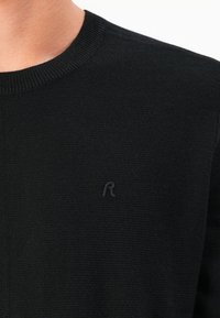 Pulover negru tricotat cu un decolteu clasic la baza gâtului și un mic logo brodat pe piept. Textură moale și un design aderent.