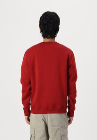 Punainen crewneck-huppari, jossa on ribbaus hihansuissa ja helmassa, ja takana pieni neliönmuotoinen märkki. Valmistettu pehmeästä, teksturoidusta materiaalista.