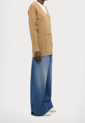 Personne portant un cardigan beige boutonné avec une ceinture chaîne dorée, un jean bleu à jambes larges et des chaussures blanches, debout devant un fond uni.