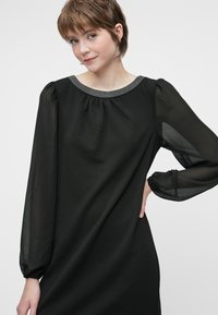 Young woman with short brown hair wearing a black dress with sheer long sleeves and a beaded neckline, posing with one hand on hip.