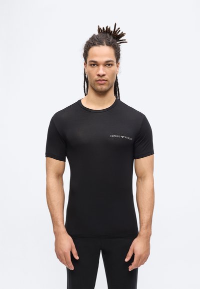 Άνδρας με ντραντλοκς που φοράει στενή μαύρη μπλούζα Emporio Armani και μαύρο παντελόνι, στέκεται μπροστά σε ένα απλό ανοιχτόχρωμο φόντο.