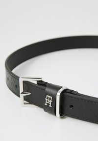 Crni kožni remen s teksturiranom površinom, sadrži srebrnu metalnu kopču i logotip pri kopči.