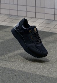 Schwarze Sneakers aus Mesh- und glatten Materialien, mit runder Zehenform, strukturiertem Sohle und goldfarbenen Hardware-Akzenten an den Schnürsenkeln und der Seite.