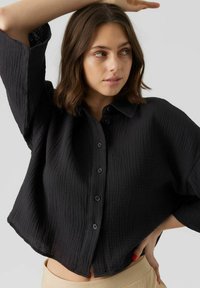 Schwarze knopflose Hemdbluse aus strukturiertem Stoff, mit Kragen, fallenden Schultern und verkürztem Saum. Akzentuiert mit schwarzen Knöpfen.