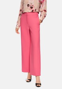 Rosa weit geschnittene Hosen aus glattem Material, mit seitlichen Taschen und hoher Taille, kombiniert mit einer floral bedruckten Bluse mit langen Ärmeln und schwarzen High Heels.