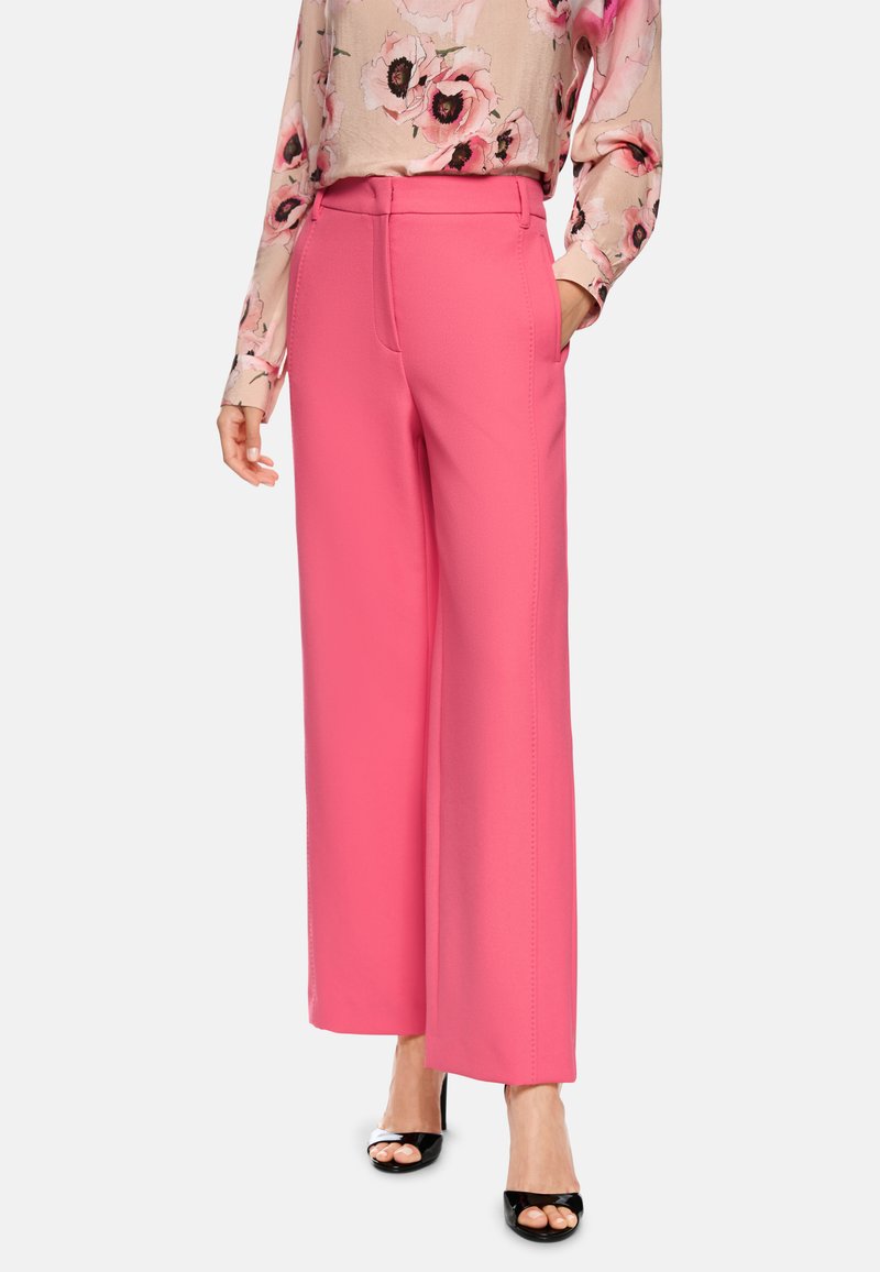 Rosa weit geschnittene Hosen aus glattem Material, mit seitlichen Taschen und hoher Taille, kombiniert mit einer floral bedruckten Bluse mit langen Ärmeln und schwarzen High Heels.