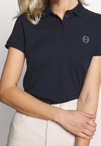 En kvinna med blont hår har på sig en mörkblå polo med en logotyp, stående med armarna i kors mot en ljus bakgrund.