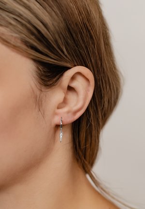 Silberne Creolen mit einem spitzen, dekorativen Anhänger und einem kleinen, klaren Edelstein-Akzent. Das Design ist schlank und minimalistisch.