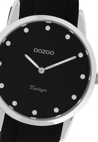 Montre noire avec un cadran rond, accents argentés et 12 marqueurs d'heure en pierres précieuses. Présente un design simple et un bracelet noir. Marque : OOZOO.