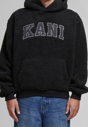 Persoon die een zwarte hoodie met structuur draagt, met "KANI" in grote grijze letters over de borst en een blauwe spijkerbroek, getoond van nek tot heupen.
