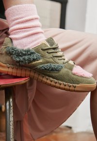 Olivgrüner Wildledersneaker mit pinkfarbenen und türkisfarbenen, flauschigen Akzenten sowie Gummisohle. Strukturiertes Obermaterial mit geschichteten Patches. Getragen mit umgeschlagenen pinken Socken.
