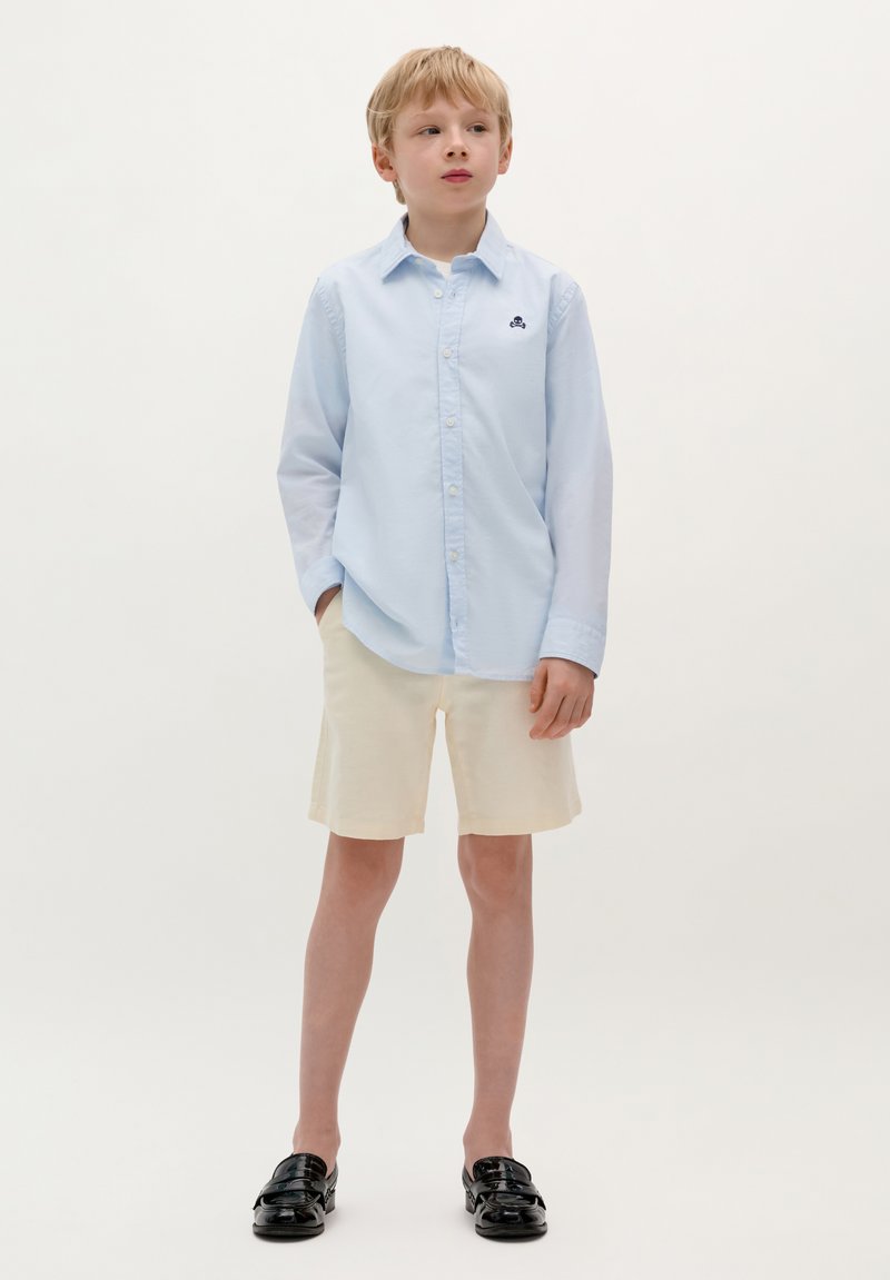 Camicia azzurro chiaro con bottoni, pantaloni corti color crema, mocassini neri. La camicia presenta un piccolo design ricamato; il tessuto sembra liscio.