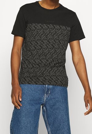 Mand iført sort Calvin Klein T-shirt med gentagende logo-mønster og blå jeans, stående mod en almindelig lys baggrund.