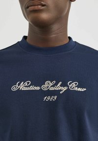 T-shirt in cotone blu navy con testo ricamato bianco: "Nautica Sailing Crew 1983." Collo a giro e maniche corte.