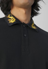 Personne portant un polo noir avec un col orné de motifs de dragons jaunes, boutons fermés près du cou, et cheveux mi-longs foncés.