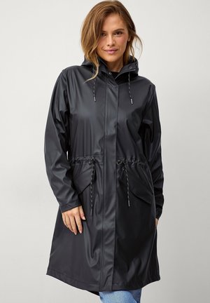 Kvinde iført en sort vandtæt hættejakke med snøre i taljen og store forlommer, stående mod en ensartet lys baggrund.