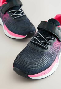 Baskets de sport noires et roses avec tige en mesh, design texturé, sangle réglable et lacets réfléchissants. Semelle blanche incurvée et semelle en caoutchouc.
