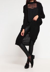 Dlouhý černý cardigan s otevřeným předním dílem, navrstvený přes top se zdobením z krajky, spárovaný s černými koženými kalhotami a plochými černými botami.