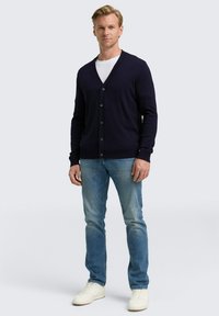 Blonder Mann, der eine schwarze Knopfleiste-Cardigan über einem weißen T-Shirt, blue Jeans und weißen Sneakers trägt und vor einem schlichten Hintergrund steht.