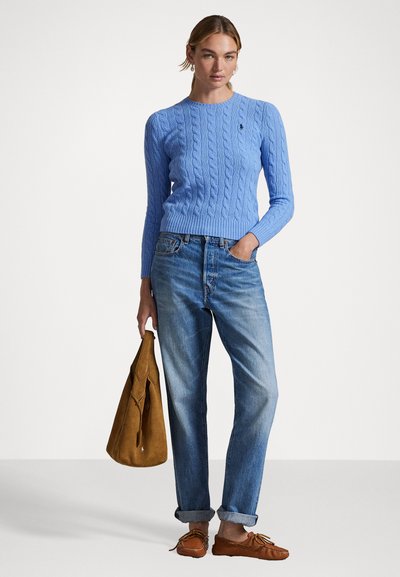 Šviesiai mėlynas megztinis su pintine tekstūra, apvaliu iškirpimu, derinamas su šviesiai mėlynomis džinsais ir rudais mokasinais. Modelis laiko rudą zomšinį krepšį.