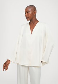 Top blanco, liso, de manga larga con escote en V y corte holgado. Hecho de una tela ligera, con detalles suaves de caída. Combinado con pantalones a juego.