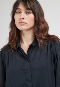 Camicia nera con bottoni realizzata in un tessuto morbido e trasparente. Presenta un colletto e lievi arricciature sulle spalle.