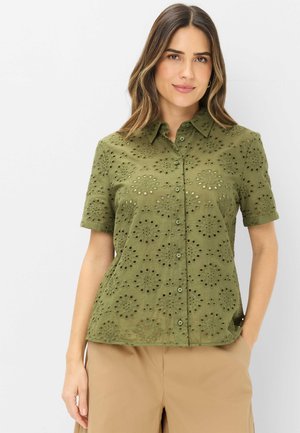 Femme portant une chemise kaki à manches courtes à boutons avec motif œillet floral et un pantalon beige, debout devant un fond blanc.